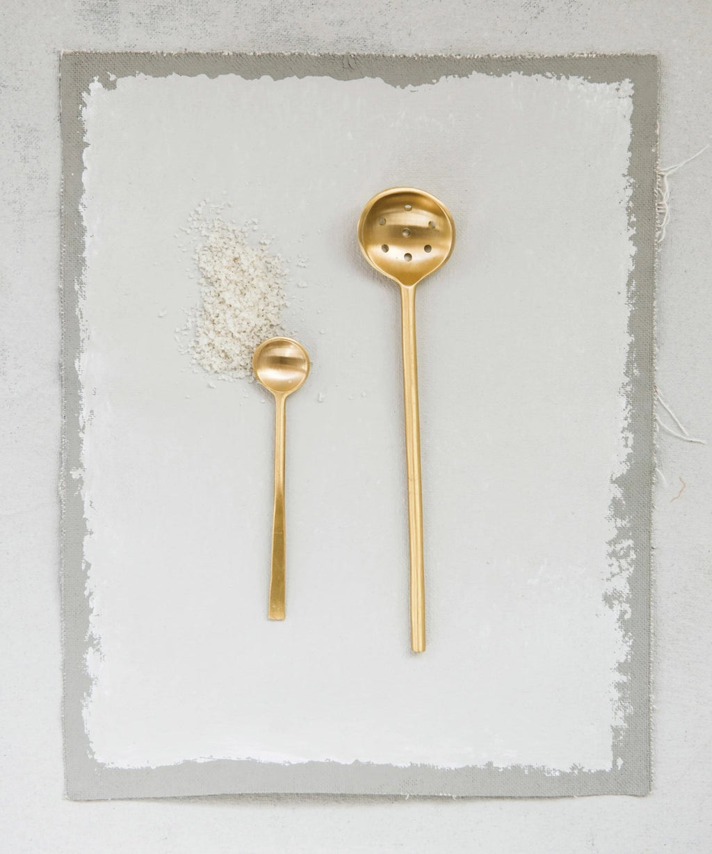 A minimalist brass slotted spoon with a long handle and round, perforated ladle styled with a small brass spoon, evoking elegance and simplicity. | Wildwood Home Co.