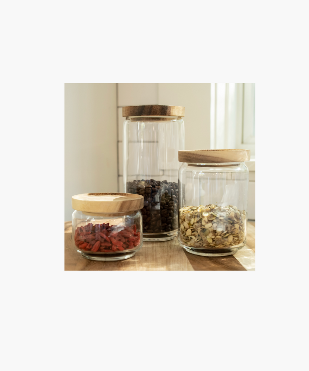 Three clear glass jars in various sizes each with a round, smooth wooden lid styled on a kitchen wooden kitchen counter. Each glass jar is filed with different dried food items. The simple, functional design conveys a minimalist aesthetic. | SW Home