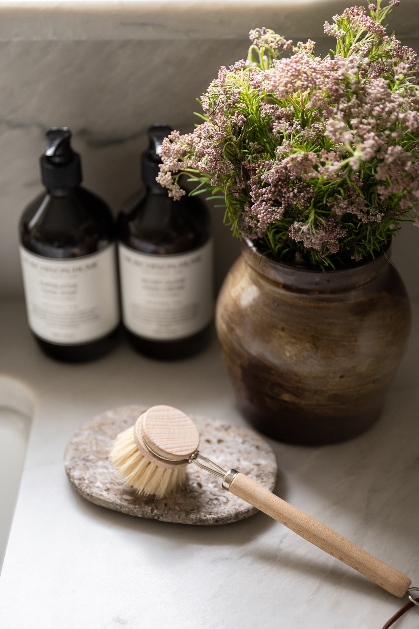 Oval shaped travertine soap dish with a rough, textured surface in shades of gray and hints of brown styled with kitchen wares. It conveys a rustic, natural, and minimalist aesthetic. | Wildwood Home Co.